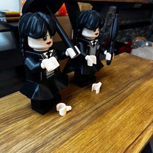 May include: Two black Lego figures of Wednesday Addams, one holding a black umbrella, the other a black object. Both figures wear black dresses with white collars and have black hair. White Lego accessories are on the wooden surface.
