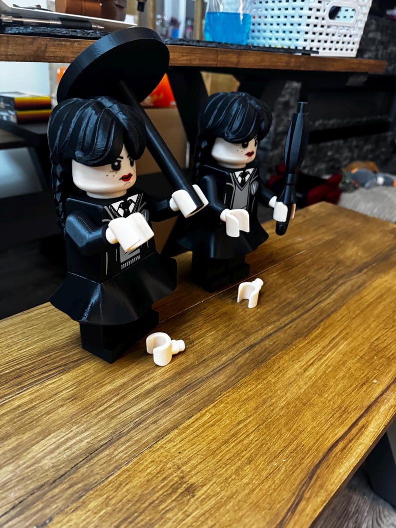 May include: Two black Lego figures of Wednesday Addams, one holding a black umbrella, the other a black object. Both figures wear black dresses with white collars and have black hair. White Lego accessories are on the wooden surface.