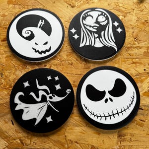 May include: Four black and white circular coasters featuring characters from The Nightmare Before Christmas. Designs include Jack Skellington, Sally, Zero, and a pumpkin face. The coasters are arranged on a wooden surface.