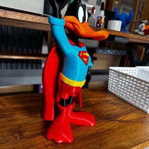 May include: A 3D-printed Daffy Duck figure in a Superman outfit. The duck is black, orange, red, blue, and yellow, with a red cape and the Superman logo on the chest. The duck is raising its left hand.