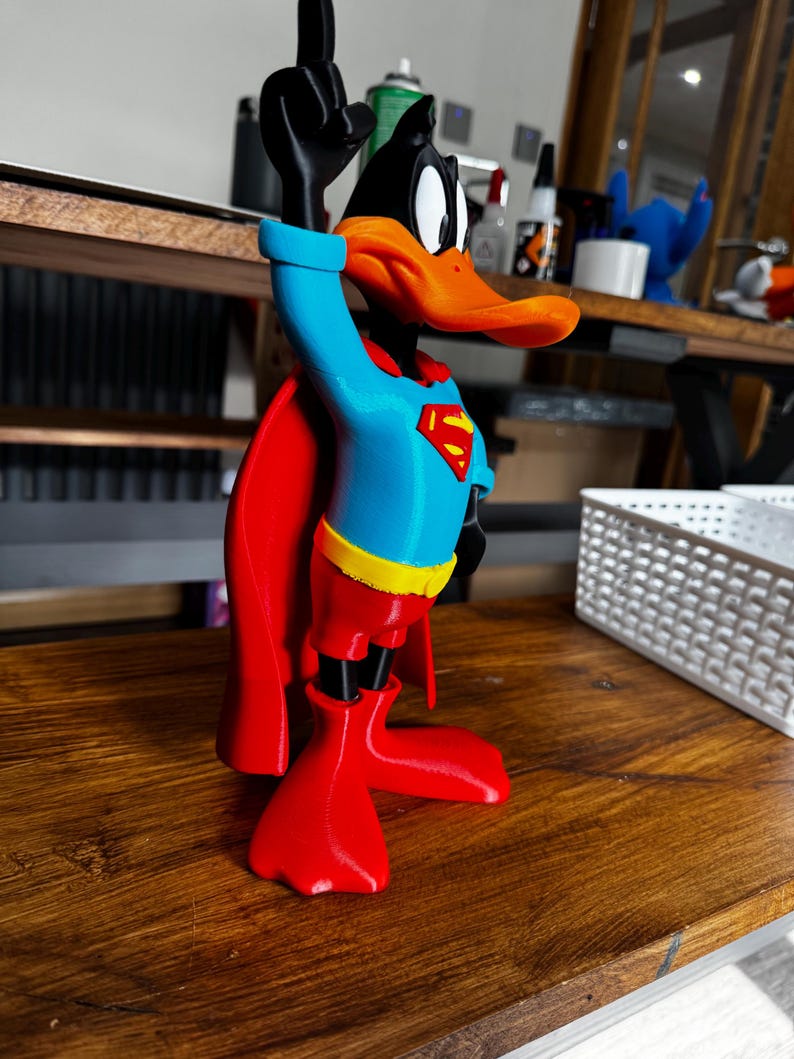 May include: A 3D-printed Daffy Duck figure in a Superman outfit. The duck is black, orange, red, blue, and yellow, with a red cape and the Superman logo on the chest. The duck is raising its left hand.
