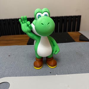 May include: A green and white Yoshi figurine with brown shoes and yellow soles. The figure is waving with its left hand. The figurine is a character from the Super Mario video game series.