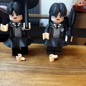May include: Two black Lego-style figures of a character with black hair, wearing a black dress, gray sweater, and white collared shirt. One figure holds a black umbrella, the other holds a black umbrella. The figures have a stern expression and red lipstick.