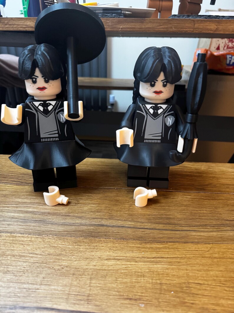 May include: Two black Lego-style figures of a character with black hair, wearing a black dress, gray sweater, and white collared shirt. One figure holds a black umbrella, the other holds a black umbrella. The figures have a stern expression and red lipstick.