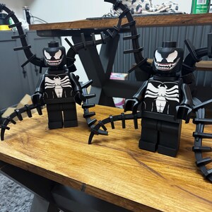 May include: Two black Lego figures of the Venom character, featuring white spider-like chest emblems and white-toothed grins. Each figure has black, spiky appendages extending from the back. They are displayed on a wooden surface.