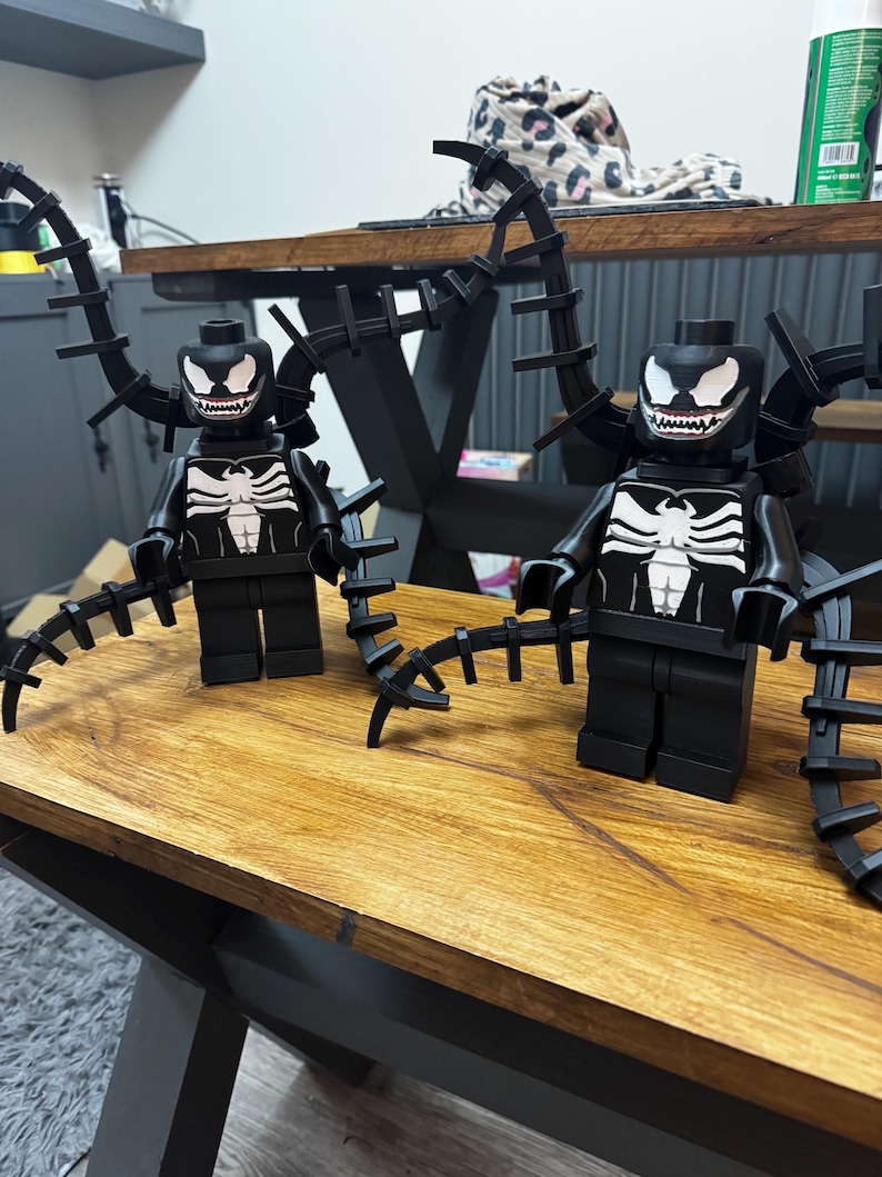 May include: Two black Lego figures of the Venom character, featuring white spider-like chest emblems and white-toothed grins. Each figure has black, spiky appendages extending from the back. They are displayed on a wooden surface.