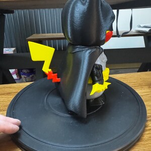 May include: A black figurine combining Batman and Pikachu, with a yellow lightning bolt tail. The figure has a black cape and bat ears, with yellow and red accents. It is on a black circular base.