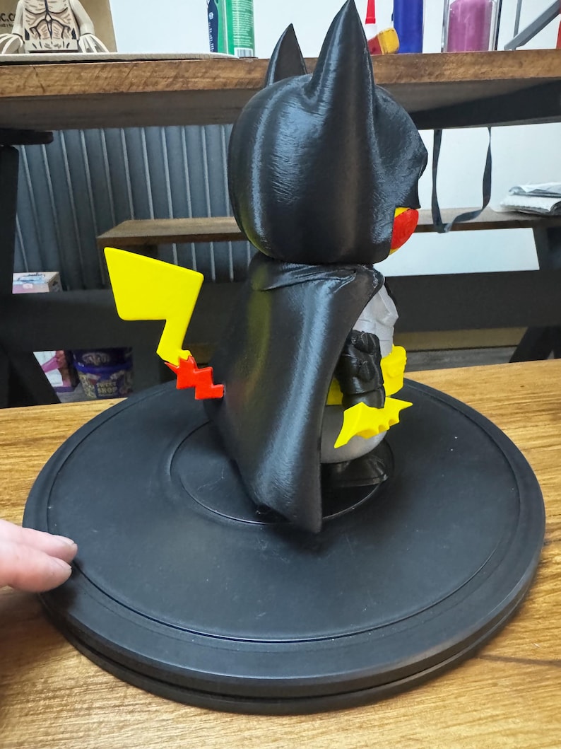May include: A black figurine combining Batman and Pikachu, with a yellow lightning bolt tail. The figure has a black cape and bat ears, with yellow and red accents. It is on a black circular base.