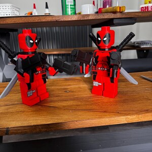 May include: Two red and black Deadpool Lego figures with black guns and silver swords. The figures have black eyes and are standing on a wooden surface. The figures are in a playful pose.