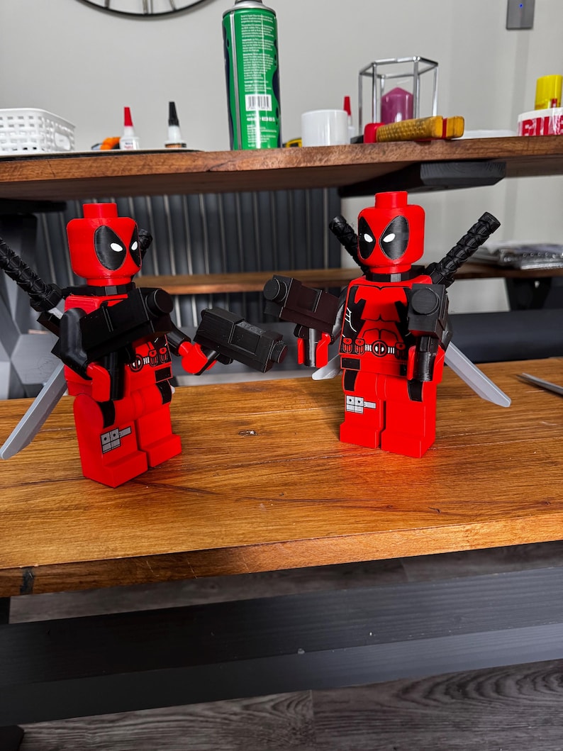 May include: Two red and black Deadpool Lego figures with black guns and silver swords. The figures have black eyes and are standing on a wooden surface. The figures are in a playful pose.