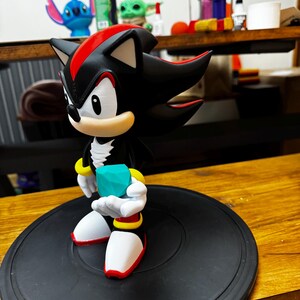 May include: A black and red figurine of Shadow the Hedgehog holding a teal gem. The figure has white gloves, red and yellow accents, and is on a black circular base. The figurine is a collectible item.