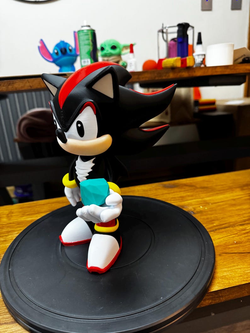 May include: A black and red figurine of Shadow the Hedgehog holding a teal gem. The figure has white gloves, red and yellow accents, and is on a black circular base. The figurine is a collectible item.