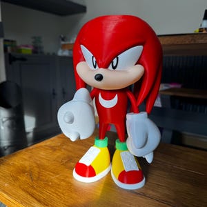 May include: A 3D-printed figure of Knuckles the Echidna, a character from the Sonic the Hedgehog franchise. The figure is primarily red with white gloves, a tan muzzle, and yellow, red, and green shoes. The figure is standing on a wooden surface.