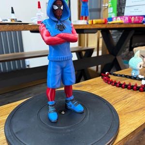May include: A 3D-printed Spider-Man figurine in a blue hoodie and shorts, red arms and legs, and blue sneakers. The figure is standing on a black circular base. Another similar figure is visible in the background.