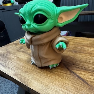 May include: A green figurine of a character with large ears and black eyes, wearing a tan robe. The figurine is standing on a wooden surface. The character's skin is a vibrant green, and the robe has a textured appearance.