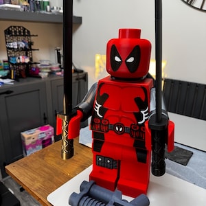 May include: A large red Deadpool figure with black accents, two black swords, and a gray gun. The figure has a detailed design, including a belt with a Deadpool logo. The swords have gold handles. The figure is a collectible item.