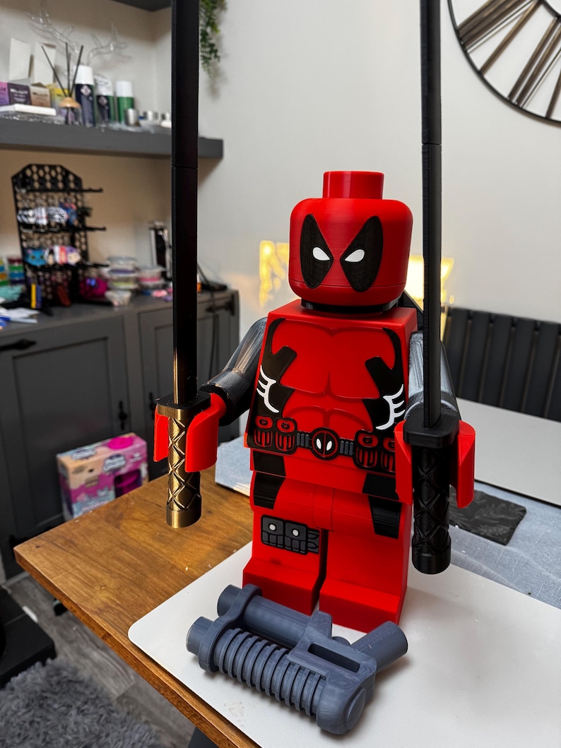 May include: A large red Deadpool figure with black accents, two black swords, and a gray gun. The figure has a detailed design, including a belt with a Deadpool logo. The swords have gold handles. The figure is a collectible item.