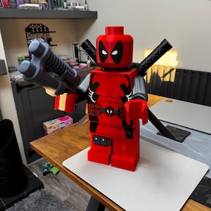 May include: A large red and black Deadpool figure, constructed from interlocking blocks, holding a gray gun and two black swords. The figure has a detailed design, including a belt and mask. The figure is displayed on a wooden table.