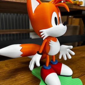 Puede incluir: Una figura impresa en 3D de Tails de la serie Sonic the Hedgehog. La figura presenta pelaje naranja y blanco, guantes blancos, zapatos rojos y blancos con detalles azules y una base verde. La figura est&aacute; en posici&oacute;n de pie.