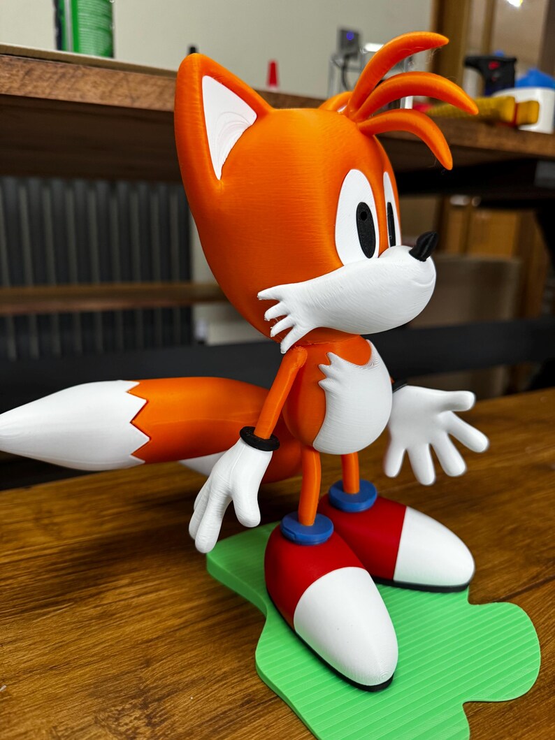 Puede incluir: Una figura impresa en 3D de Tails de la serie Sonic the Hedgehog. La figura presenta pelaje naranja y blanco, guantes blancos, zapatos rojos y blancos con detalles azules y una base verde. La figura est&aacute; en posici&oacute;n de pie.