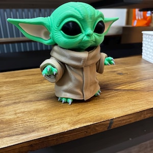 May include: A green figurine of a character with large ears and black eyes, wearing a tan robe. The figurine is on a wooden surface. The character's hands and feet have green claws. The figurine is a popular character from a science fiction franchise.
