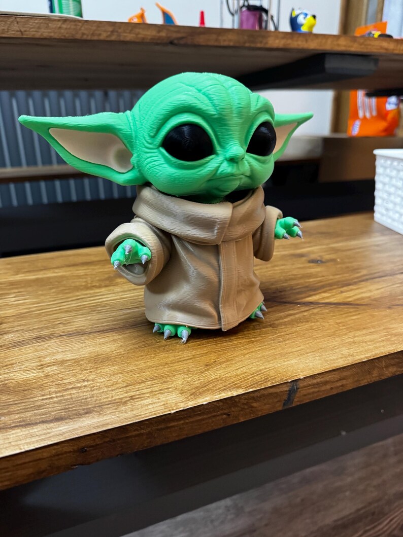 May include: A green figurine of a character with large ears and black eyes, wearing a tan robe. The figurine is on a wooden surface. The character's hands and feet have green claws. The figurine is a popular character from a science fiction franchise.
