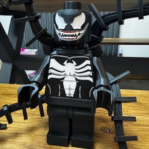 May include: A black Venom-themed Lego figure with white accents. The figure features a detailed face with a wide grin and a white spider emblem on the chest. Additional black appendages extend from the figure's back.
