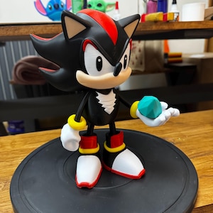 May include: A black and red Shadow the Hedgehog figurine holding a teal Chaos Emerald. The figure has white gloves, red and yellow accents on the shoes and wrists, and is posed on a black circular base. The figurine is a collectible item.