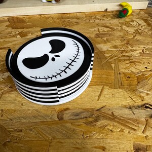 May include: A stack of black and white striped coasters with a Jack Skellington face design. The coasters are round and have a glossy finish. A small gnome figurine with a yellow hat and bee-striped body is visible in the background.