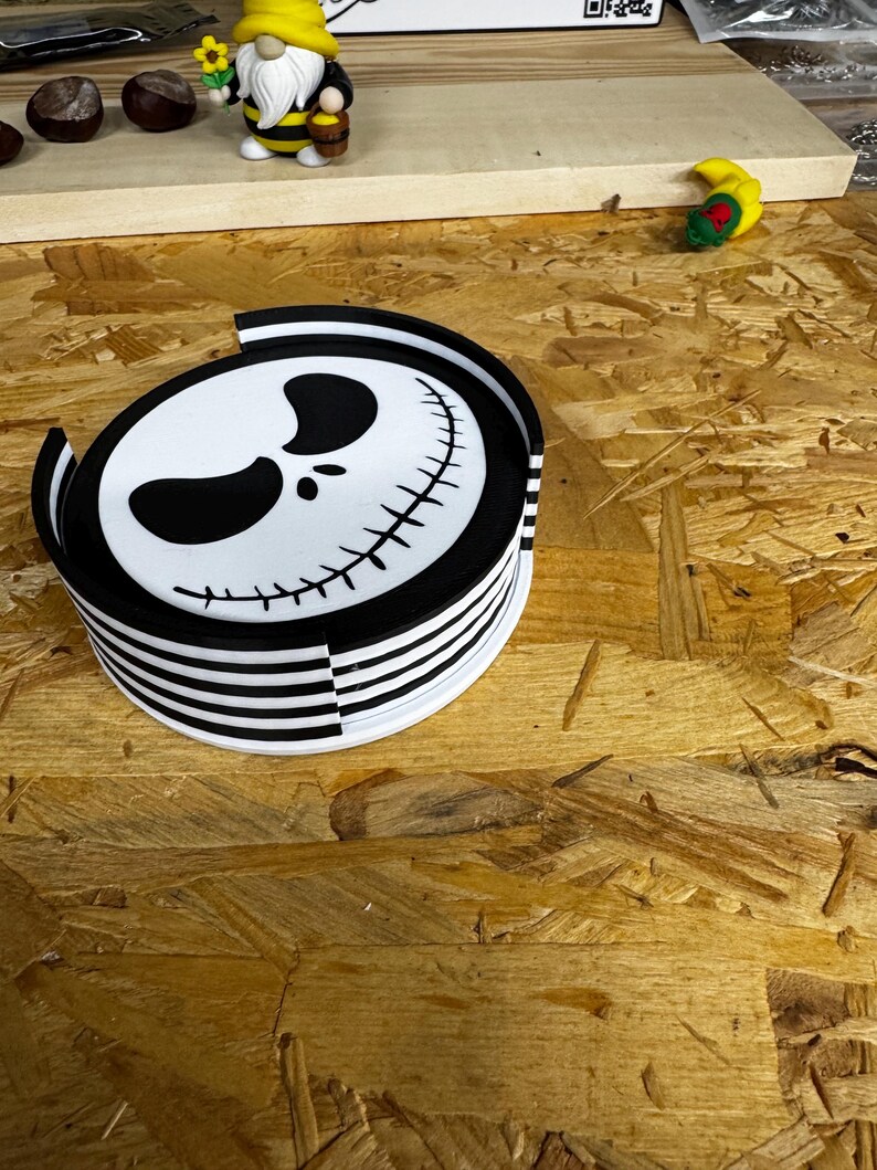 May include: A stack of black and white striped coasters with a Jack Skellington face design. The coasters are round and have a glossy finish. A small gnome figurine with a yellow hat and bee-striped body is visible in the background.