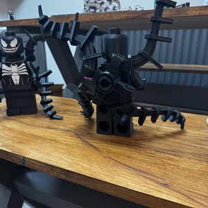 May include: Black Lego Venom figure with articulated appendages and a detailed white spider emblem on the chest. The figure has a menacing grin and is posed on a wooden surface. A second Lego figure is also visible.