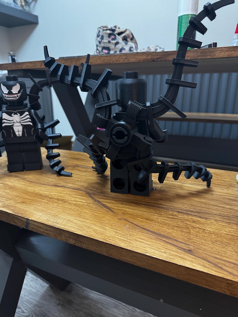 May include: Black Lego Venom figure with articulated appendages and a detailed white spider emblem on the chest. The figure has a menacing grin and is posed on a wooden surface. A second Lego figure is also visible.