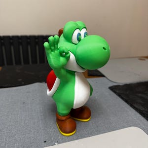 May include: A green and white Yoshi figurine with a red shell and brown shoes. The figurine is in a standing position with its right hand raised in an "okay" gesture. The figurine has large blue eyes and a long snout.