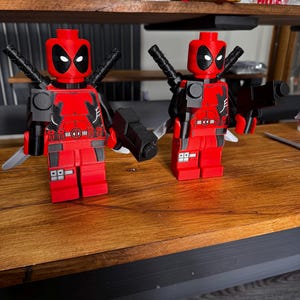 May include: Two red and black Deadpool figures, each with a black gun, katana swords, and a detailed torso design. The figures are posed on a wooden surface, showcasing the iconic mask and costume.
