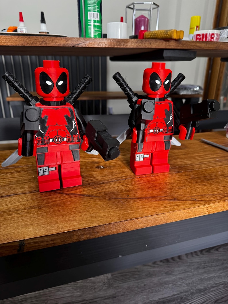 May include: Two red and black Deadpool figures, each with a black gun, katana swords, and a detailed torso design. The figures are posed on a wooden surface, showcasing the iconic mask and costume.