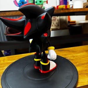 May include: A black and red figurine of a character with spiky hair, red accents, and white gloves. The figure is standing on a black circular base. The character is from the Sonic the Hedgehog franchise.