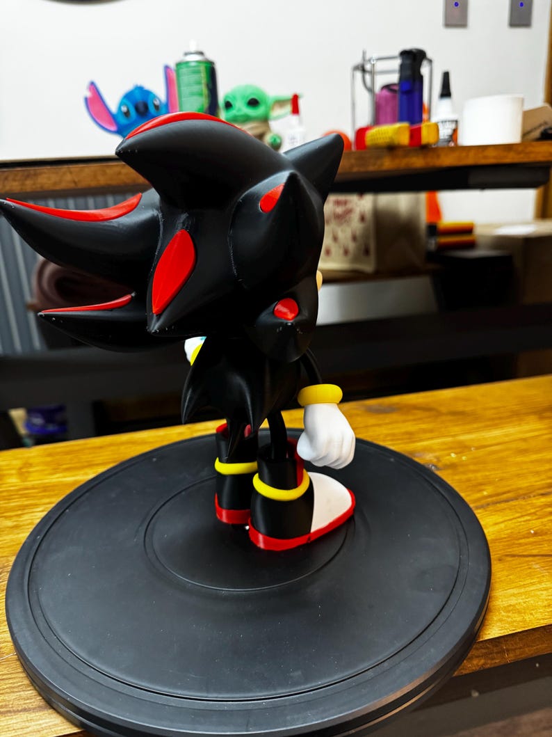 May include: A black and red figurine of a character with spiky hair, red accents, and white gloves. The figure is standing on a black circular base. The character is from the Sonic the Hedgehog franchise.