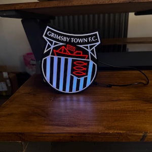 May include: A lighted Grimsby Town F.C. emblem, featuring a shield design with the team's name and a ship illustration. The emblem has a black and white striped background with blue neon lighting. The emblem is on a wooden surface.