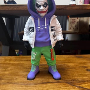 May include: A Joker action figure wearing a white jacket, purple hoodie, and green pants. The figure has green hair, white face paint, and red lipstick. The pants read "BORN TO BE BAD". The figure is wearing purple boots and black gloves.