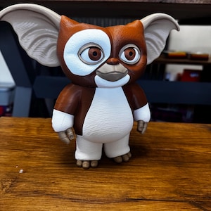 May include: A stylized figurine of the character Gizmo from Gremlins, featuring large pointed ears, brown and white fur-like texture, a white chest and face patch, and large brown eyes. The figure stands upright with a smooth, matte finish.