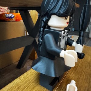 May include: A black Lego figure of a character with a black dress, black hair in braids, and a black umbrella. The figure has a white collar and a neutral skin tone. A detached white Lego hand is on the wooden surface.