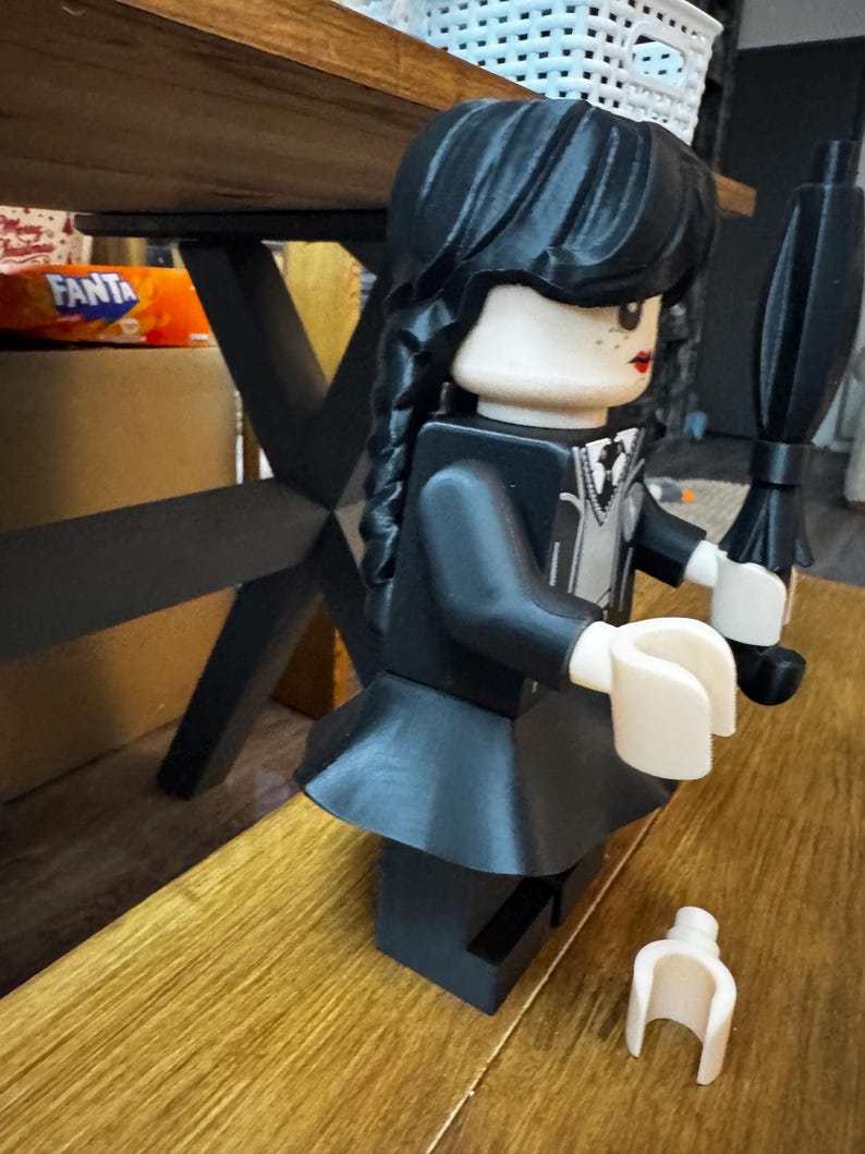 May include: A black Lego figure of a character with a black dress, black hair in braids, and a black umbrella. The figure has a white collar and a neutral skin tone. A detached white Lego hand is on the wooden surface.