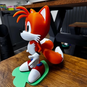 Puede incluir: Figura impresa en 3D de Tails de la serie Sonic the Hedgehog. La figura presenta pelaje naranja y blanco, zapatos azules, calcetines rojos y guantes blancos. Se encuentra sobre una base verde.