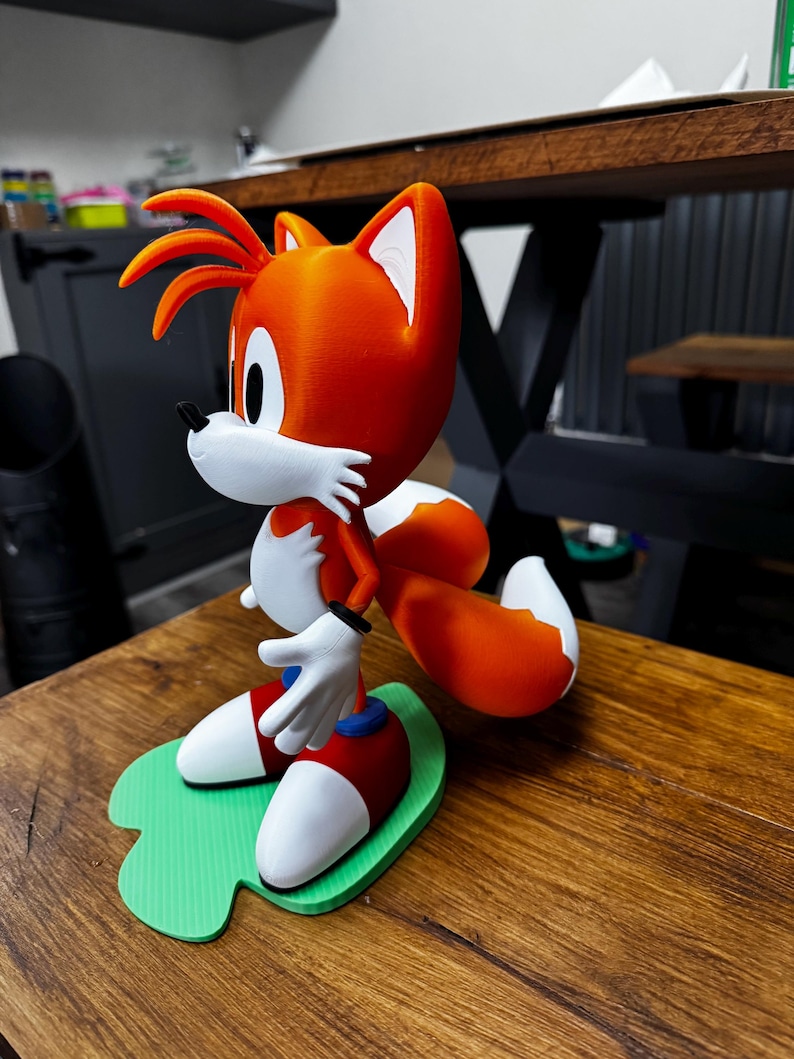 Puede incluir: Figura impresa en 3D de Tails de la serie Sonic the Hedgehog. La figura presenta pelaje naranja y blanco, zapatos azules, calcetines rojos y guantes blancos. Se encuentra sobre una base verde.