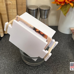 May include: A white, geometric food container with a clear glass bowl underneath. The container is held together with rubber bands. The image also includes a "Made in U.S.A." sticker.