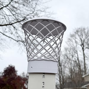 May include: A white, lattice-design chimney cap. The cap is cylindrical with a domed top and is mounted on a white pipe. The background features bare tree branches and a cloudy sky.