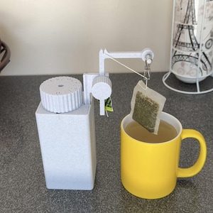 May include: A white tea bag holder with a tea bag suspended over a yellow mug filled with tea. The holder is next to a white container and a basket of fruit. A tea-themed item.