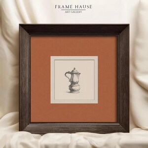 May include: A square wall art piece featuring a dark brown wooden frame with a burnt orange mat board. The center contains a cream-colored print of a silver coffee pot illustration. The text FRAME HAUSE ART GALLERY is printed at the top.