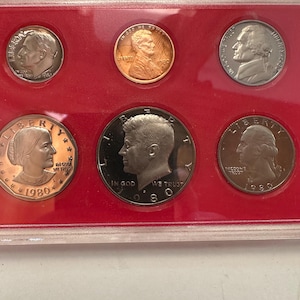 May include: A set of 1980 US coins, including a penny, nickel, dime, quarter, and half-dollar, displayed in a red plastic holder. The coins feature portraits of historical figures and the words "In God We Trust" and "Liberty".