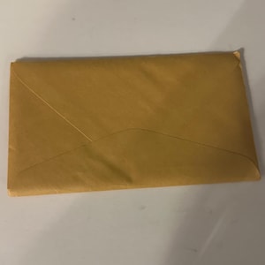 Può includere: Una busta rettangolare chiusa in carta marrone chiaro. La busta ha un lembo triangolare ed è sigillata. La carta ha un aspetto leggermente strutturato. La busta è appoggiata su una superficie bianca.
